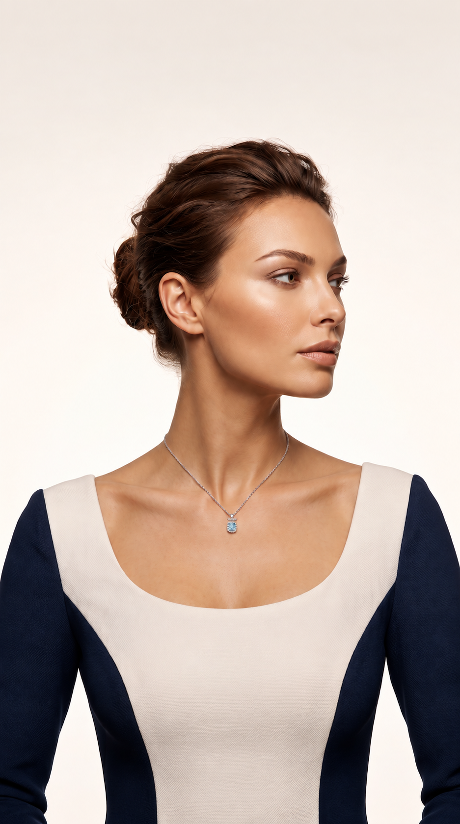A woman with her hair in a bun wears a delicate silver necklace with a small light blue pendant, paired with a color-blocked top in cream and navy blue, set against a plain light beige background.