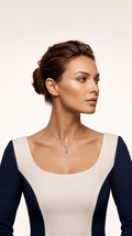 A woman with her hair in a bun wears a delicate silver necklace with a small light blue pendant, paired with a color-blocked top in cream and navy blue, set against a plain light beige background.