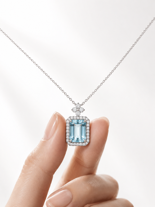 A close-up of a delicate silver necklace with a rectangular light blue gemstone pendant, held between two fingers against a bright white background.