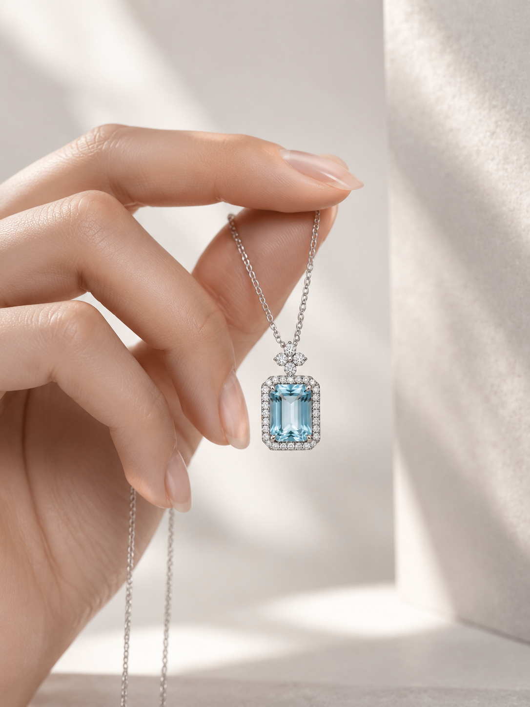 A hand holds a delicate silver necklace with a rectangular light blue gemstone pendant surrounded by small clear stones, set against a softly lit neutral background.